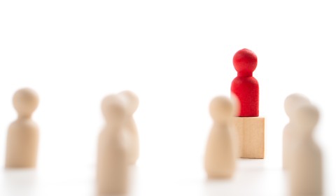 Wooden pegs representing people like those in a board game stand
      about.
      The leader peg-person is red and stands on a platform to addres the 6 
      other wood-coloured pegs.
      The photographer has used a depth of field effect to place the leader
      in sharp focus while allowing the crowd to blur, the background is
      pure white.