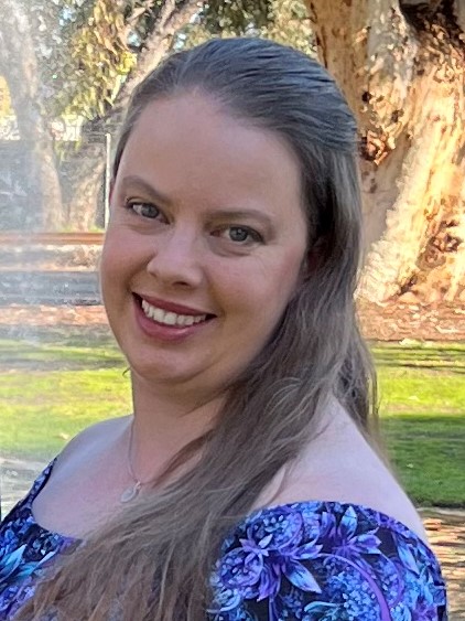 Elizabeth smiles, turning her head to look towards the camera.
      She has long brown hair and is wearing a summery blue-purple dress.